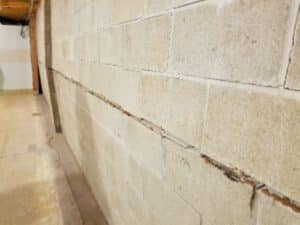 A textured concrete basement wall with a horizontal crack running through the middle, extending into a spacious, aging room with exposed pipes above.