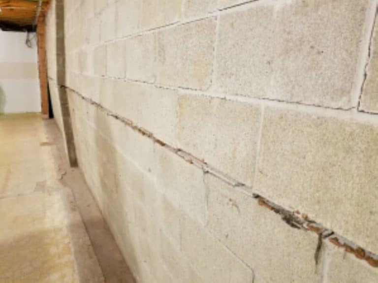 A textured concrete basement wall with a horizontal crack running through the middle, extending into a spacious, aging room with exposed pipes above.