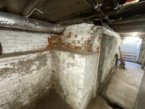 Dingy basement with exposed brick walls, peeling white paint, and visible pipes. A dimly lit, narrow passage leads to stairs. Feels old and neglected.