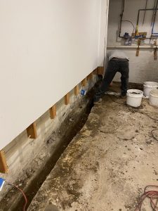 A person works in a basement, with a trench dug along a white wall. Exposed pipes and buckets are nearby, suggesting a plumbing or repair project.