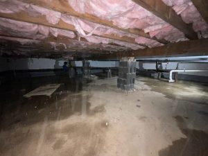 Crawl space with exposed pink insulation under wooden beams. Damp floor with pipes visible. Worker in the background suggests repair work. Gloomy atmosphere.