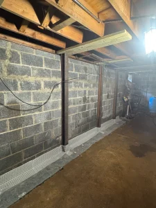 Basement under renovation with exposed brick walls, wooden beams, and new drainage installation along the floor. A bright work light illuminates the space.