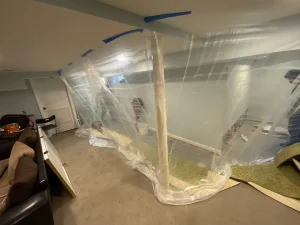 A basement under renovation, with plastic sheeting taped to the ceiling, partitioning the space. Tools and materials are visible in the area.