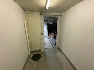Dimly lit basement corridor with white walls and exposed beam ceiling. A doorway leads to a cluttered area with stored items, conveying a utilitarian feel.