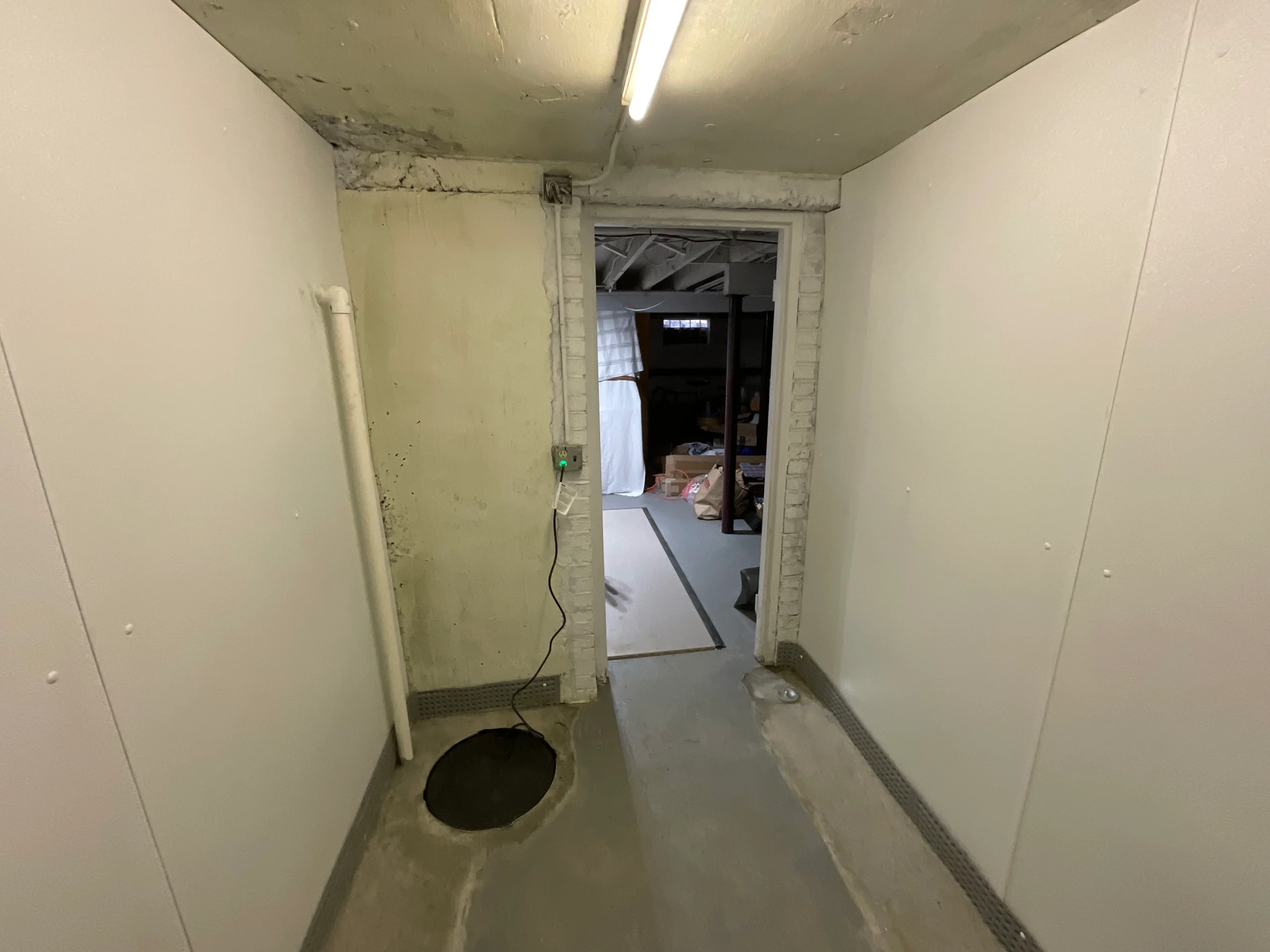 Dimly lit basement corridor with white walls and exposed beam ceiling. A doorway leads to a cluttered area with stored items, conveying a utilitarian feel.