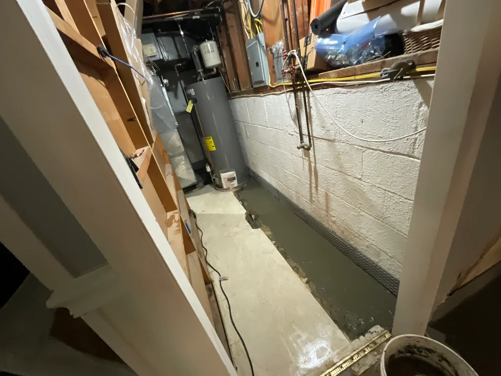 Basement with wet concrete floor, exposed brick wall, water heater, shelves, and pipes. The scene suggests ongoing maintenance or repair work.