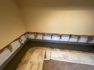 Basement under renovation with exposed wall studs and a plastic membrane. Concrete floor partially removed. Neutral-colored walls, dim lighting.