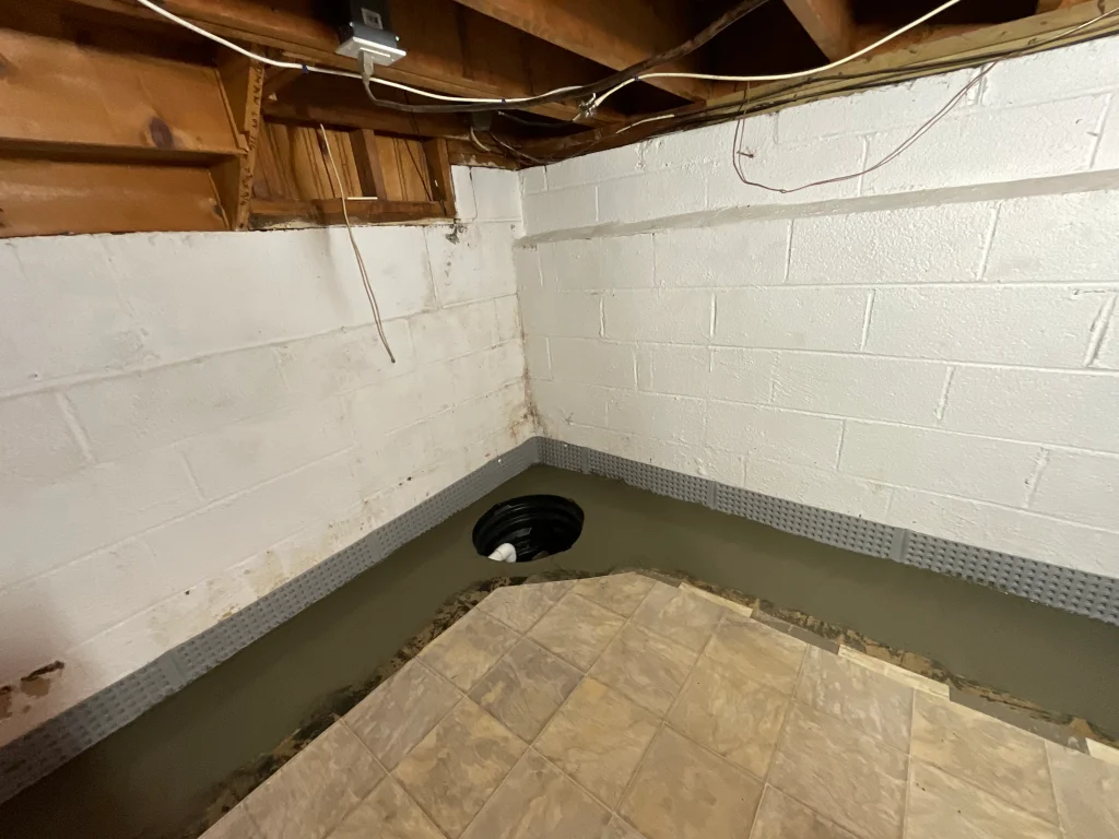 Basement corner with white cinder block walls, exposed wooden beams, and a partially finished floor. A sump pit is visible, suggesting repairs.