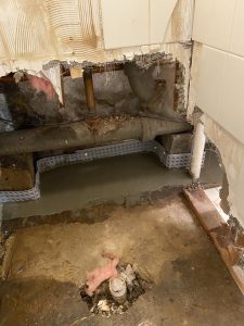 A damaged wall exposes rusted pipes and insulation in a construction area. Broken tiles and debris highlight disrepair and need for renovation.
