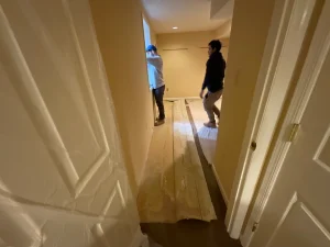 Two people are working in a narrow hallway with bare floors, one near a window and the other standing. The space is under renovation, with a focused and busy atmosphere.