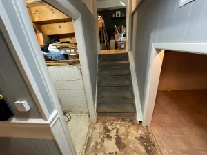 A narrow hallway with a stairway leading up to a cluttered storage area. The basement floor is worn, with visible damage. The atmosphere is cluttered and dimly lit.