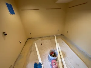 Empty yellow room with unfinished floor covered by tarp. Tools and wooden planks lie near a bucket in the center. Blue painter's tape on the wall.
