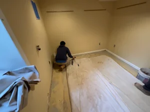 A person kneels in a bare room with yellow walls, preparing to install flooring. A large canvas is spread on the floor, with tools and materials nearby.