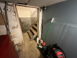 Dimly lit basement with concrete stairs leading up, flanked by rough walls. A broom, mop, and cleaning supplies are leaning against a blue-gray wall, creating a cluttered, utilitarian atmosphere.